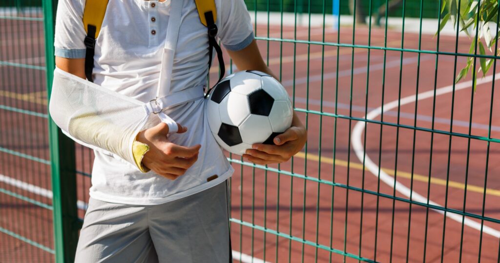 Teenager with broken arm holding football ball next to sports ground. Boy in white t shirt with hand splint in a arm sling for support and reducing pressure on neck. Injury, traumas, accident by kids